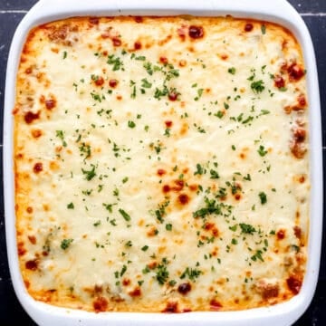 Overhead view of baked easy lasagna recipe in square white baking dish on black tile surface.