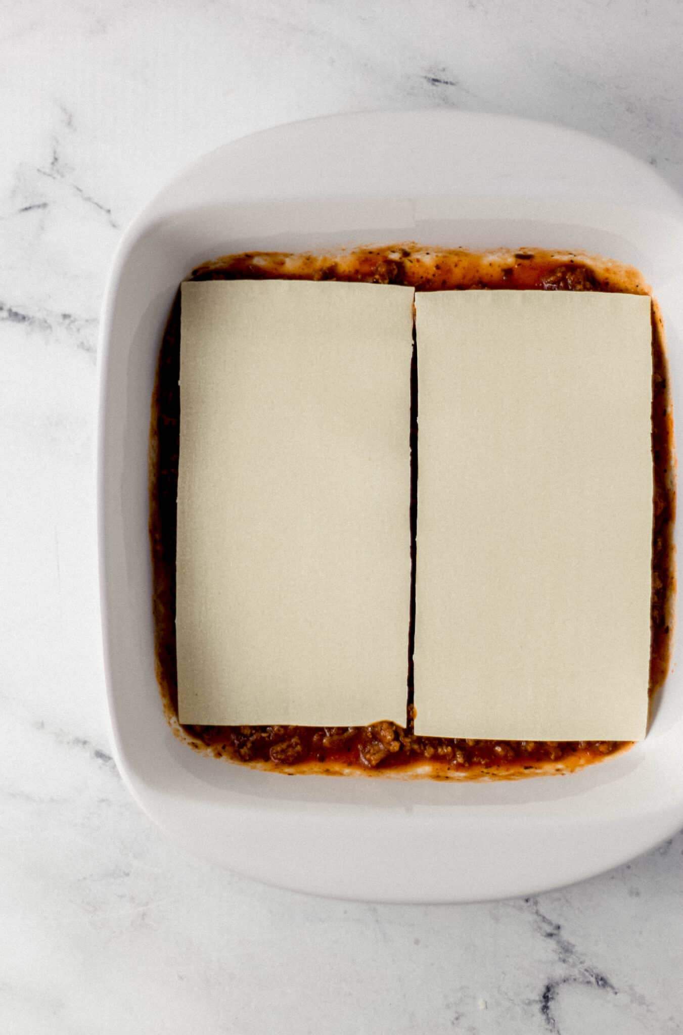 meat sauce and noodles added to white square baking dish 