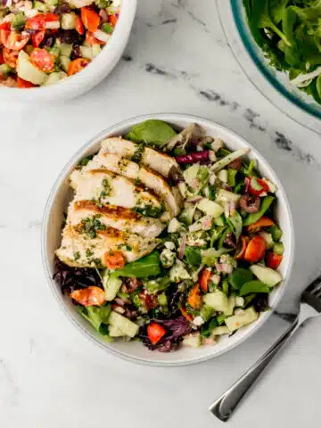 overhead view of greek chicken salad in white bowl beside a fork and two other bowls with the rest of the ingredients
