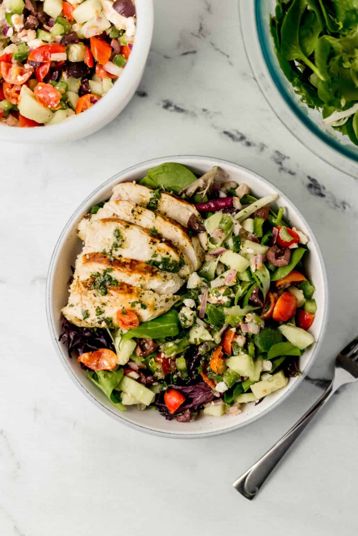 overhead view of Greek chicken salad in white bowl beside a fork, bowl of  mixed greens, and vegetable blend