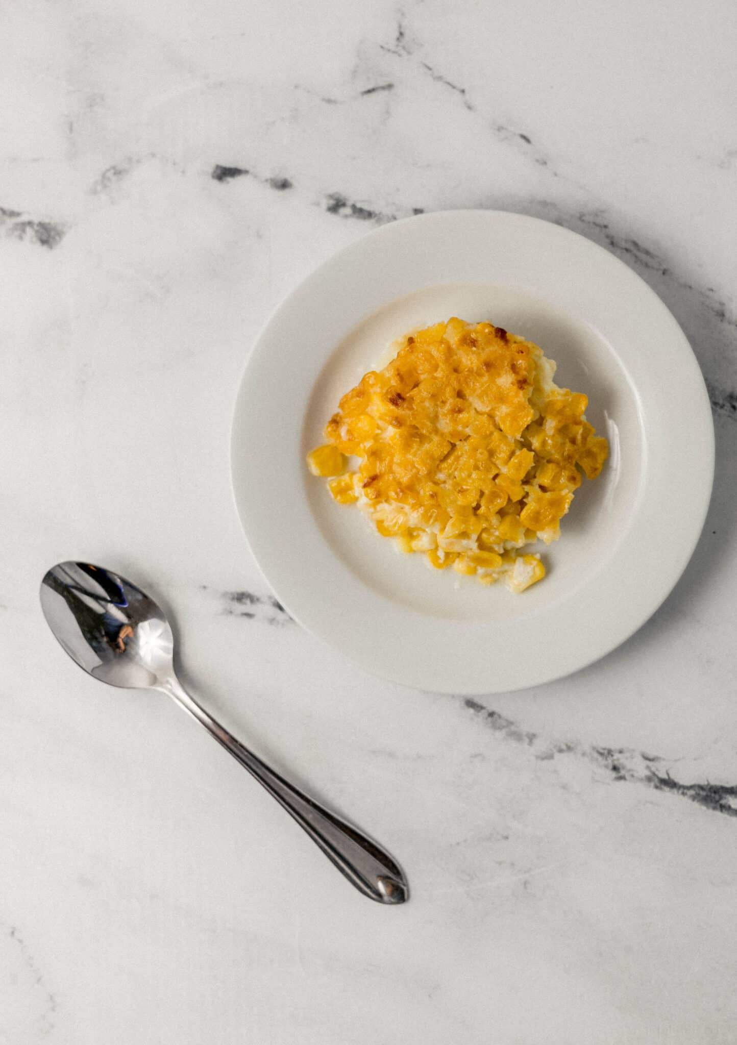 overhead view of single serving of corn pudding on small white plate beside a spoon 