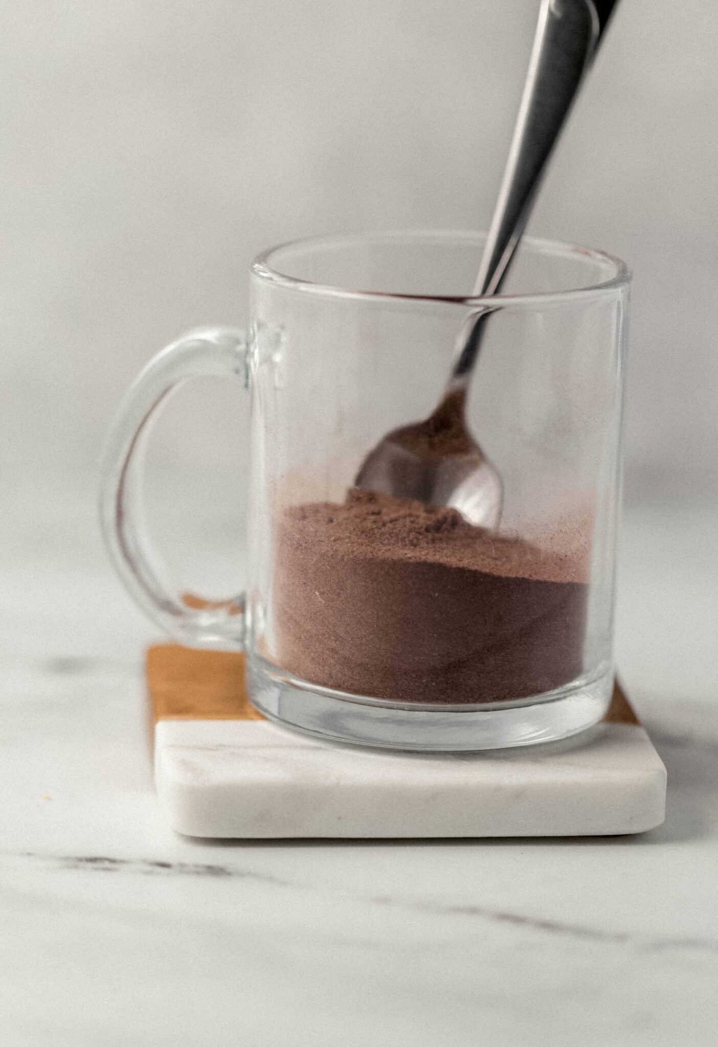 close up side view of flour, sugar, and cocoa powder added to glass mug with spoon