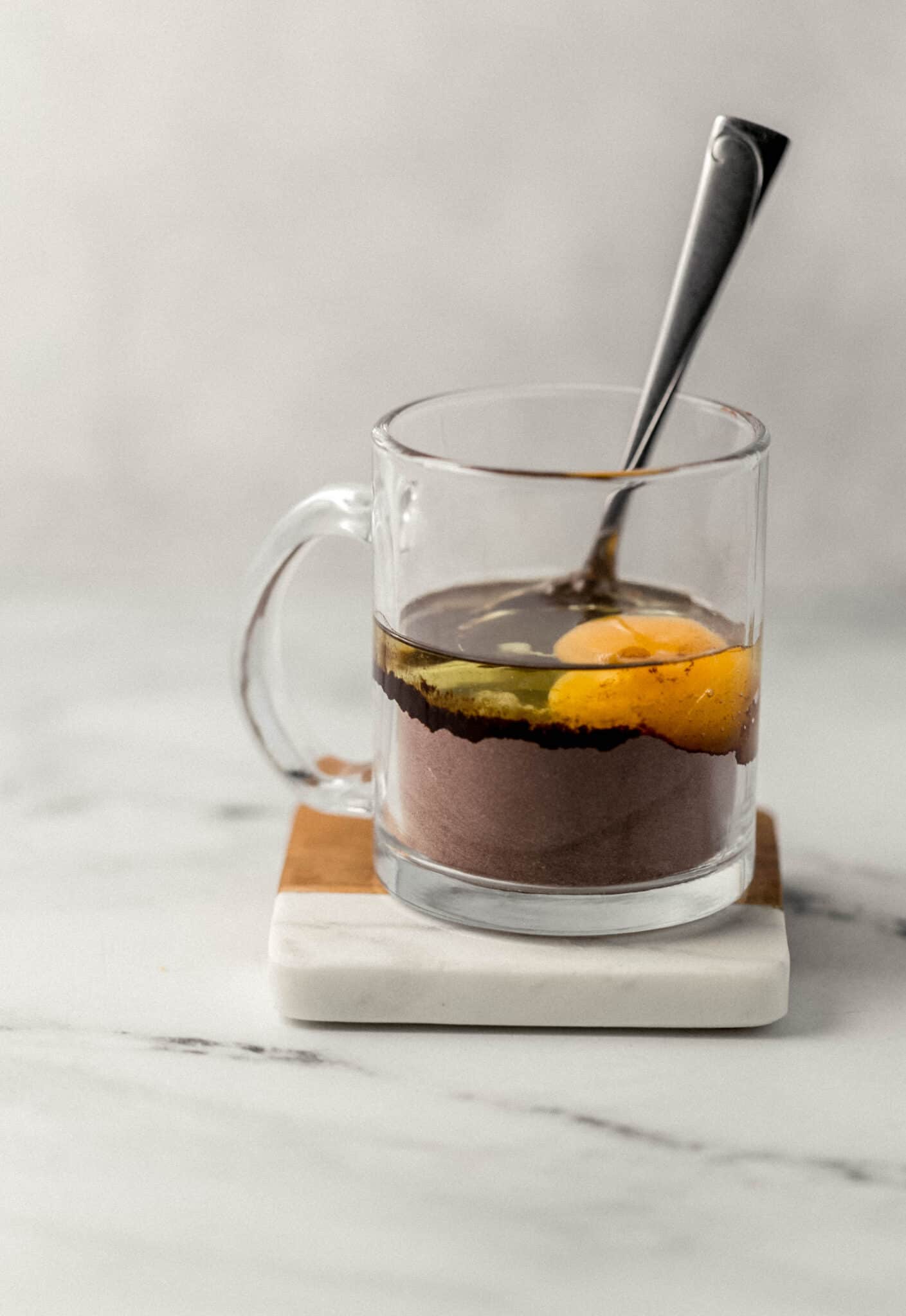 close up side view of egg and oil added to glass mug with spoon