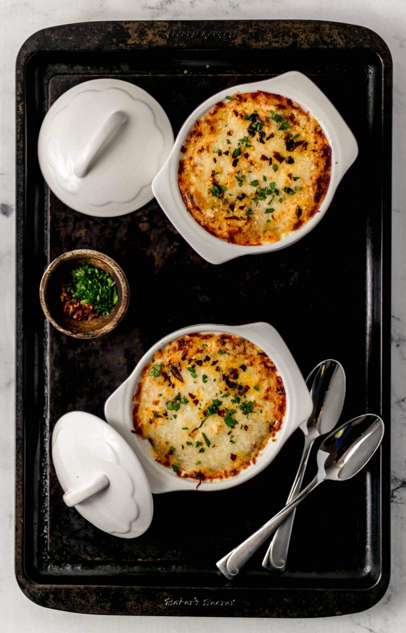 overhead view of two finished pizza bowls on sheet pan with spoons and lids to ramekins