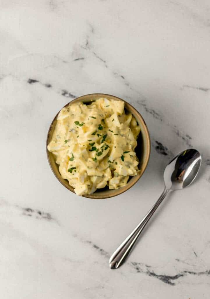 overhead view of egg salad in a bowl beside a spoon on marble surface
