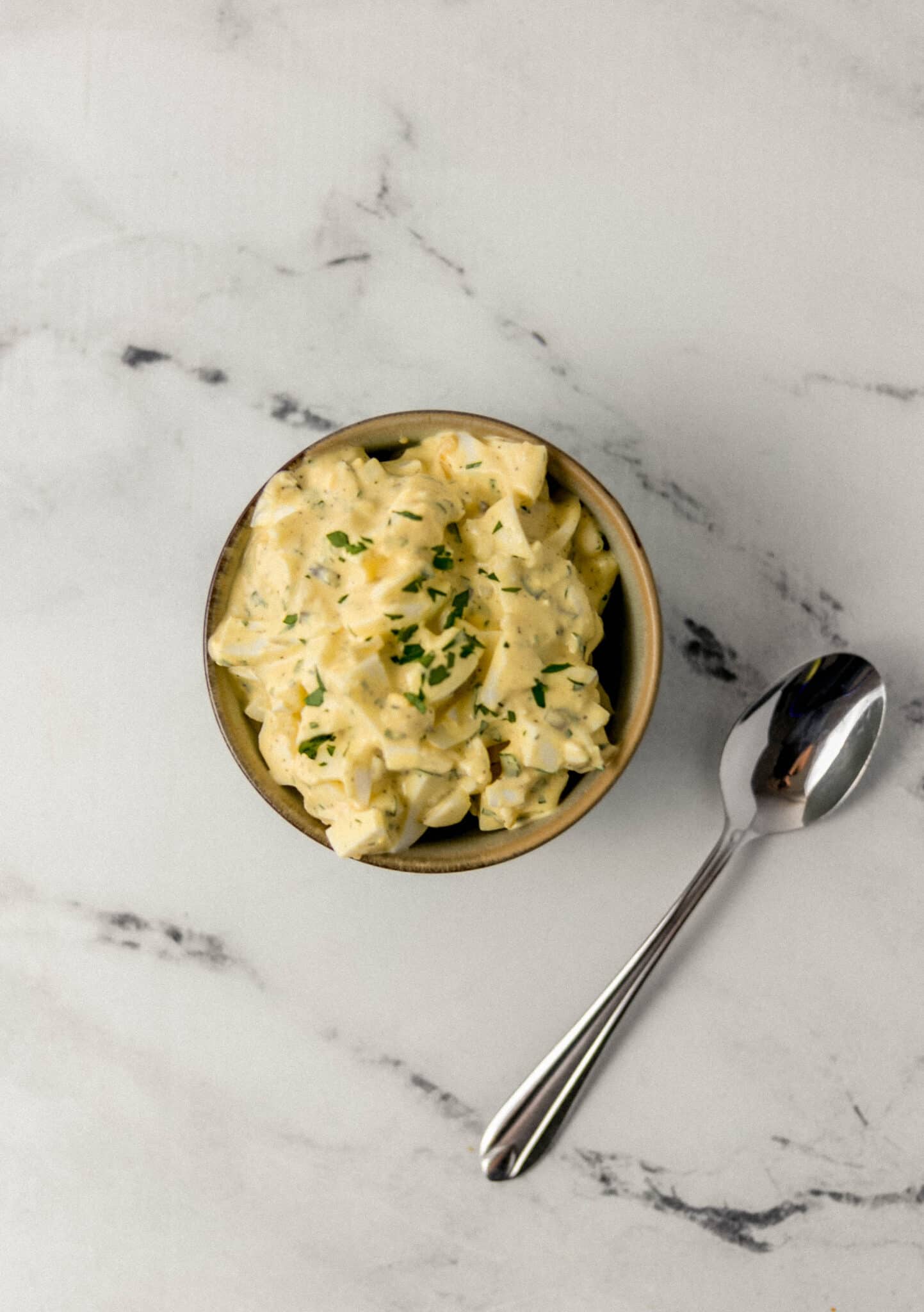 overhead view of finished egg salad in bowl beside a spoon on marble surface