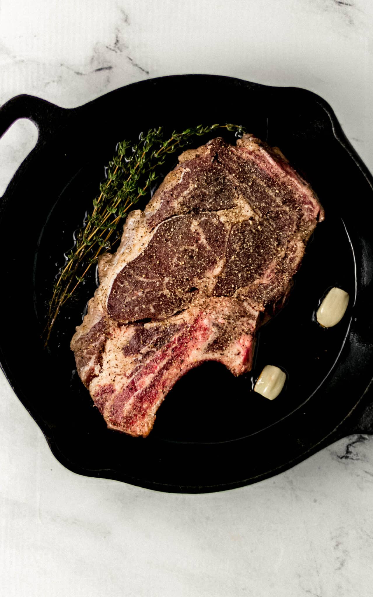 seasoned steak in cast iron skillet with hot oil, fresh thyme, and two garlic cloves.