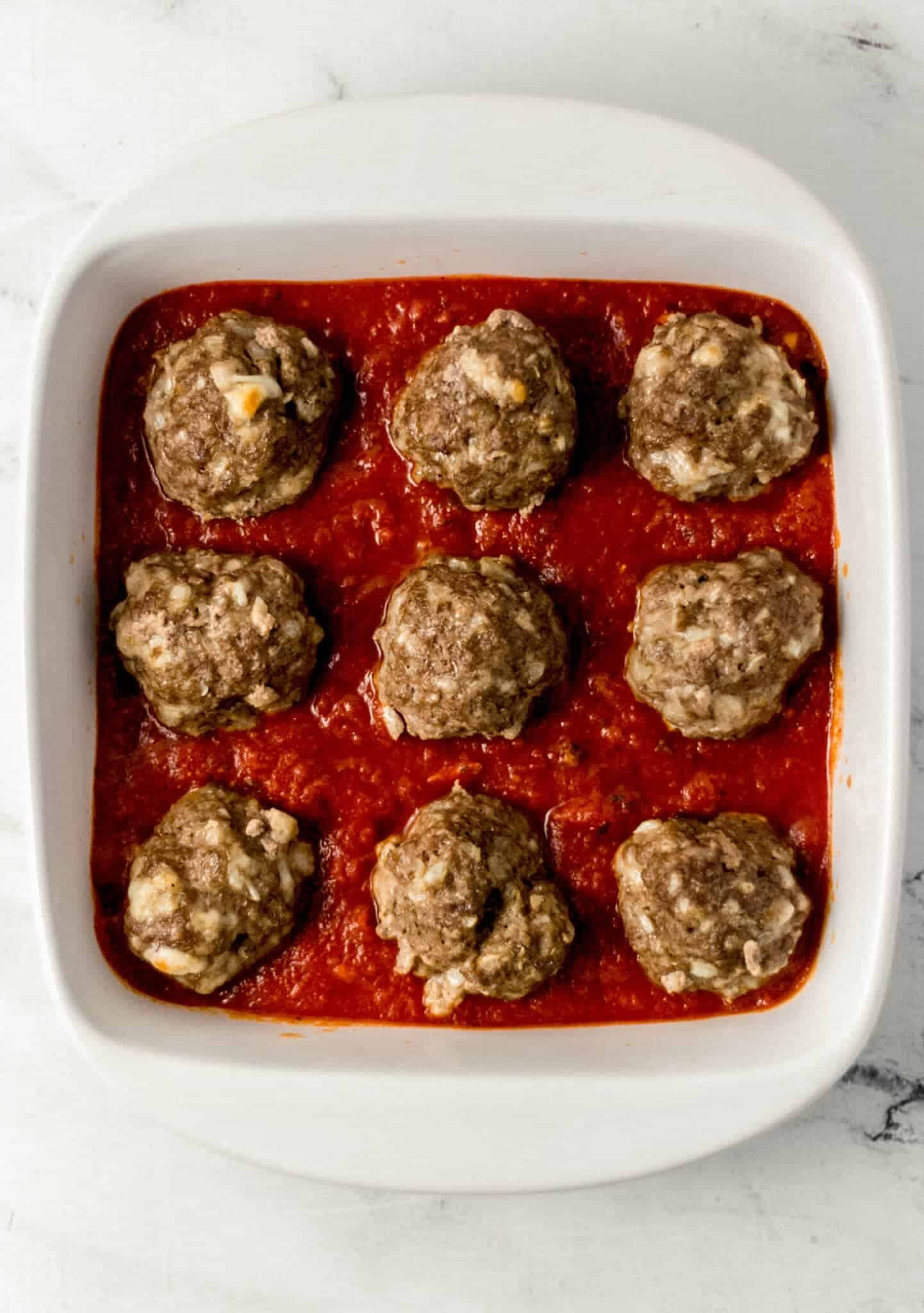 cooked meatballs place on top of pasta sauce in white baking dish