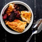 Small white bowl with single serving of blackberry cobbler in it by spoon on black tile surface.