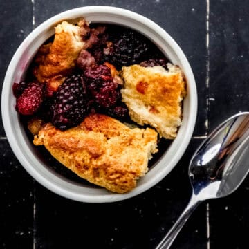 Small white bowl with single serving of blackberry cobbler in it by spoon on black tile surface.