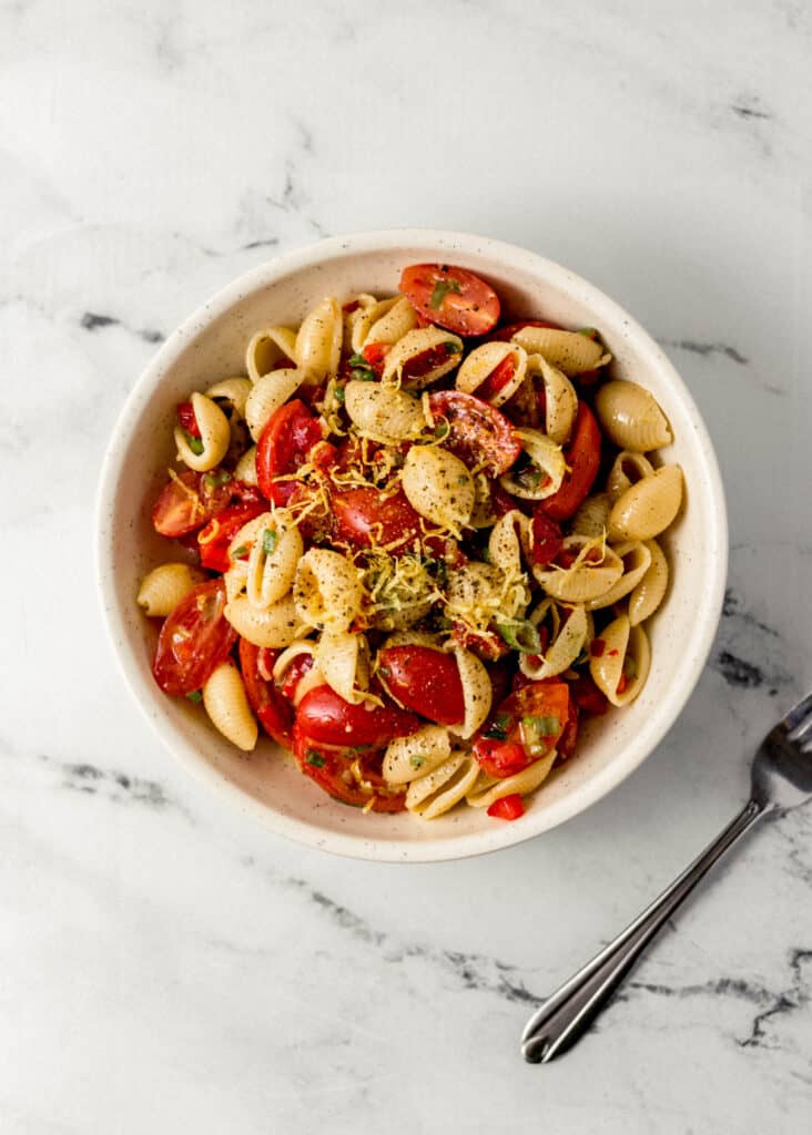 overhead view of pasta salad in a white bowl beside a fork on marble surface