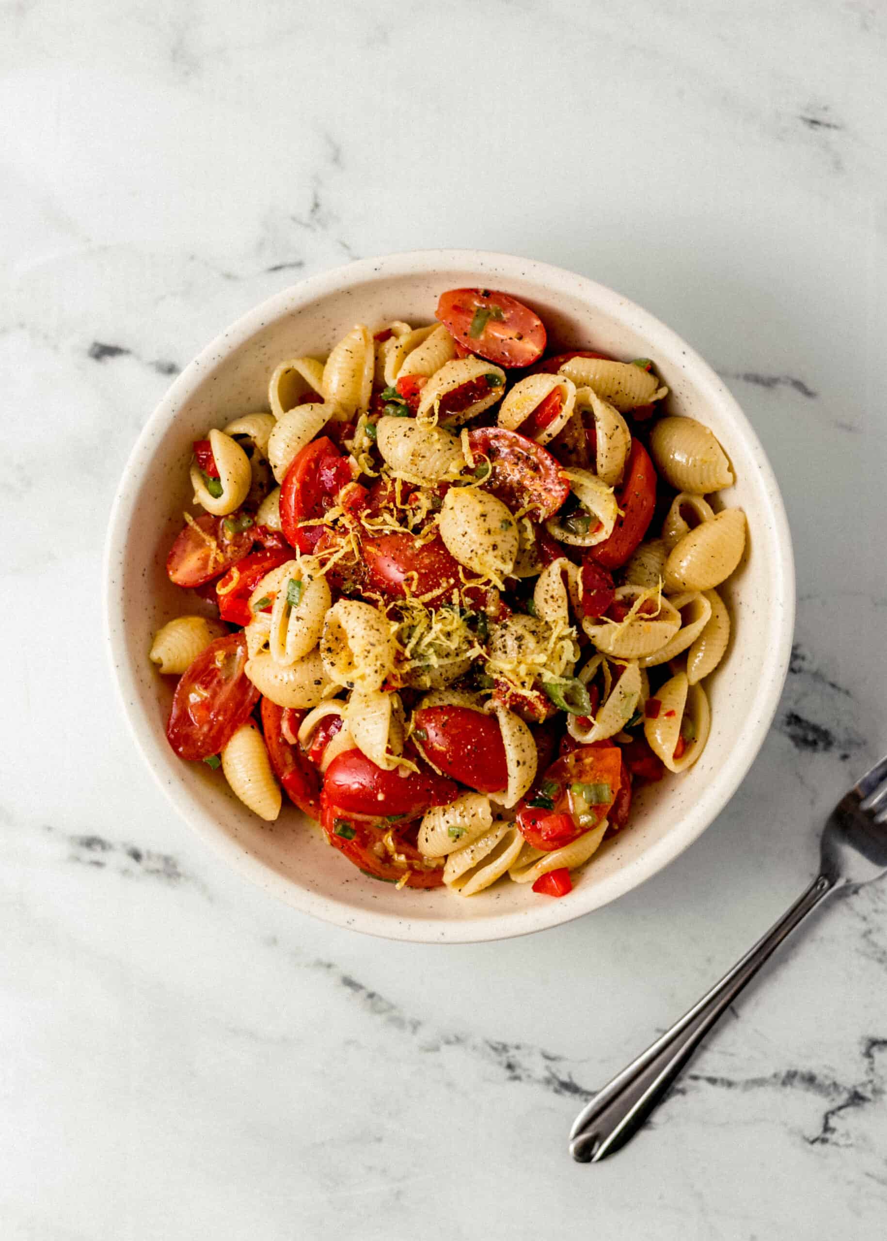 overhead view of finished pasta salad in white bowl beside a fork on marble surface 