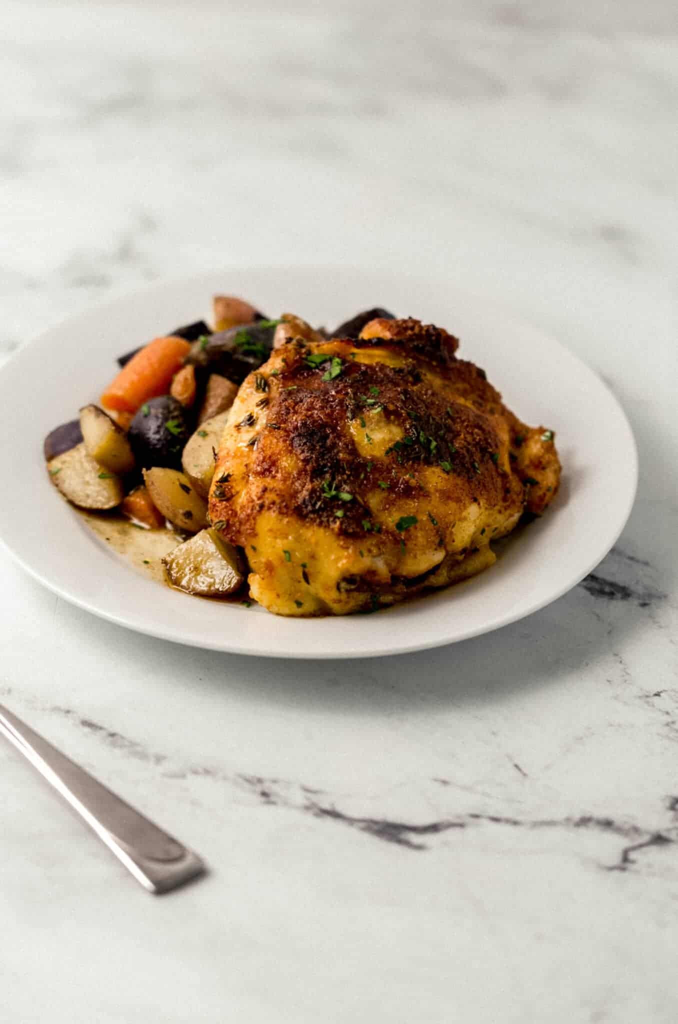 close up side view of piece of chicken thigh with serving of potatoes and carrots on white plate 