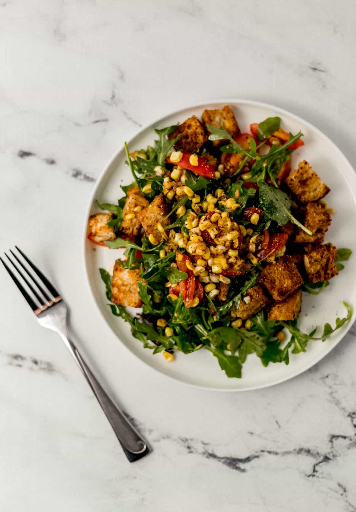 overhead view of serving of corn salad on white plate beside a fork on marble surface 