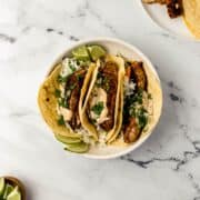 Overhead view of finished tacos in white bowl on marble surface by other ingredients.