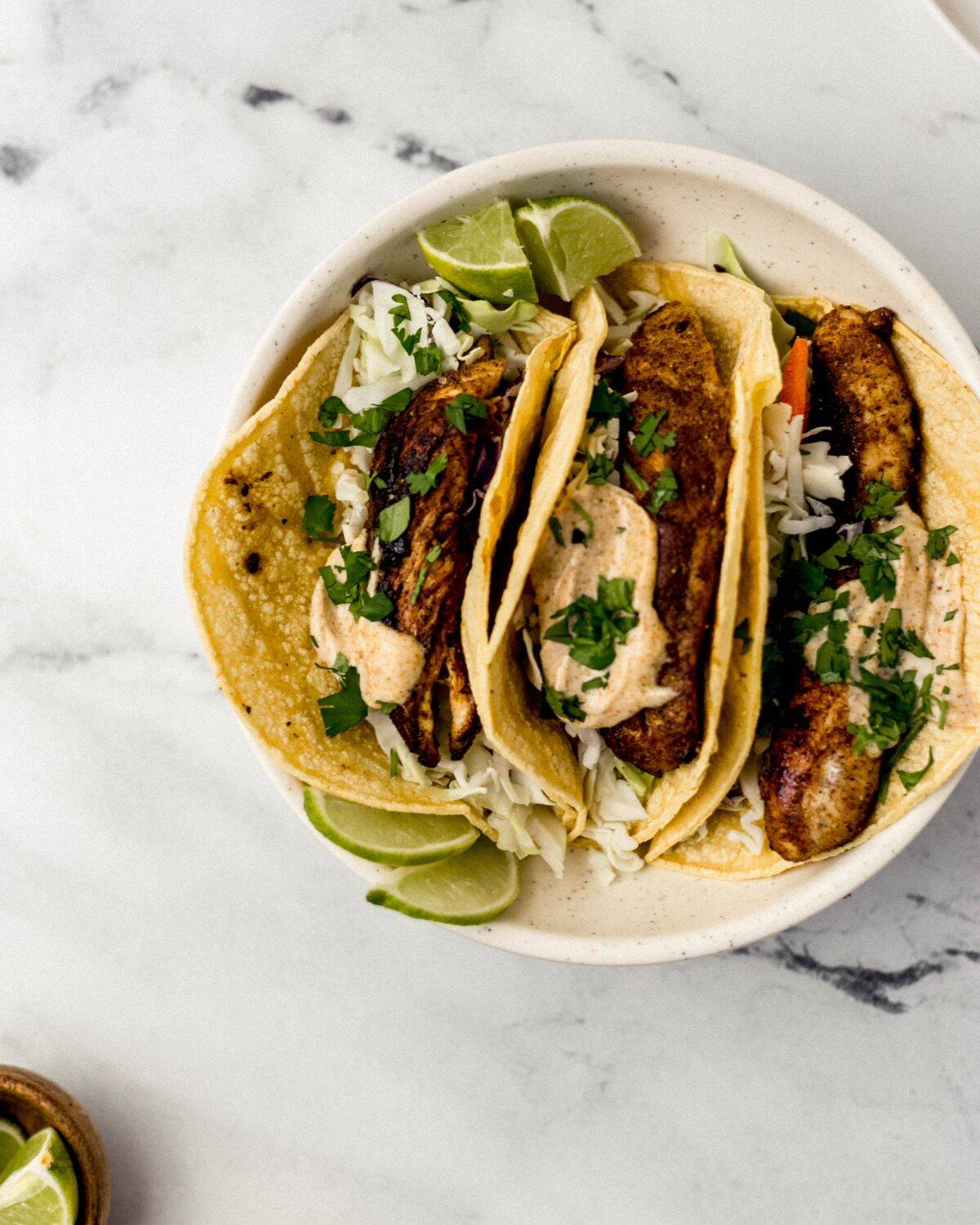 Jerk Chicken Tacos Simply LaKita 3 Overhead view of three tacos in white bowl by small wooden bowl of lime wedges.