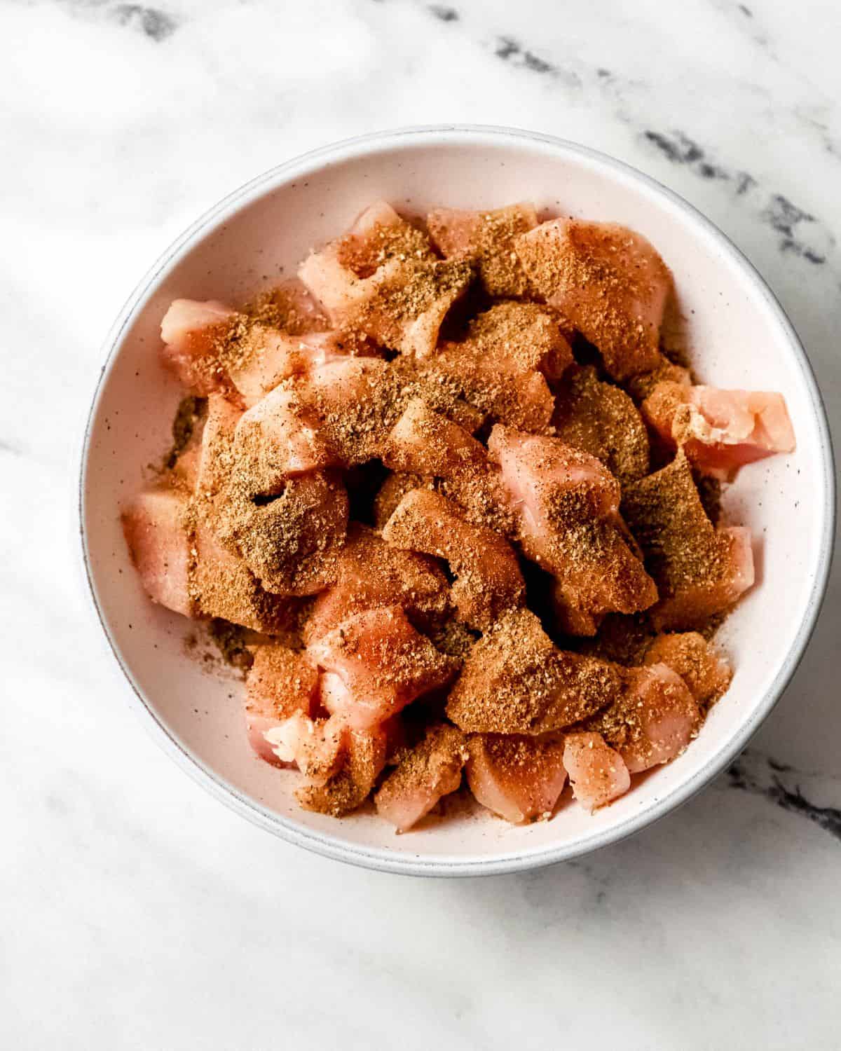 Overhead view of raw chicken topped with seasonings in white bowl. 