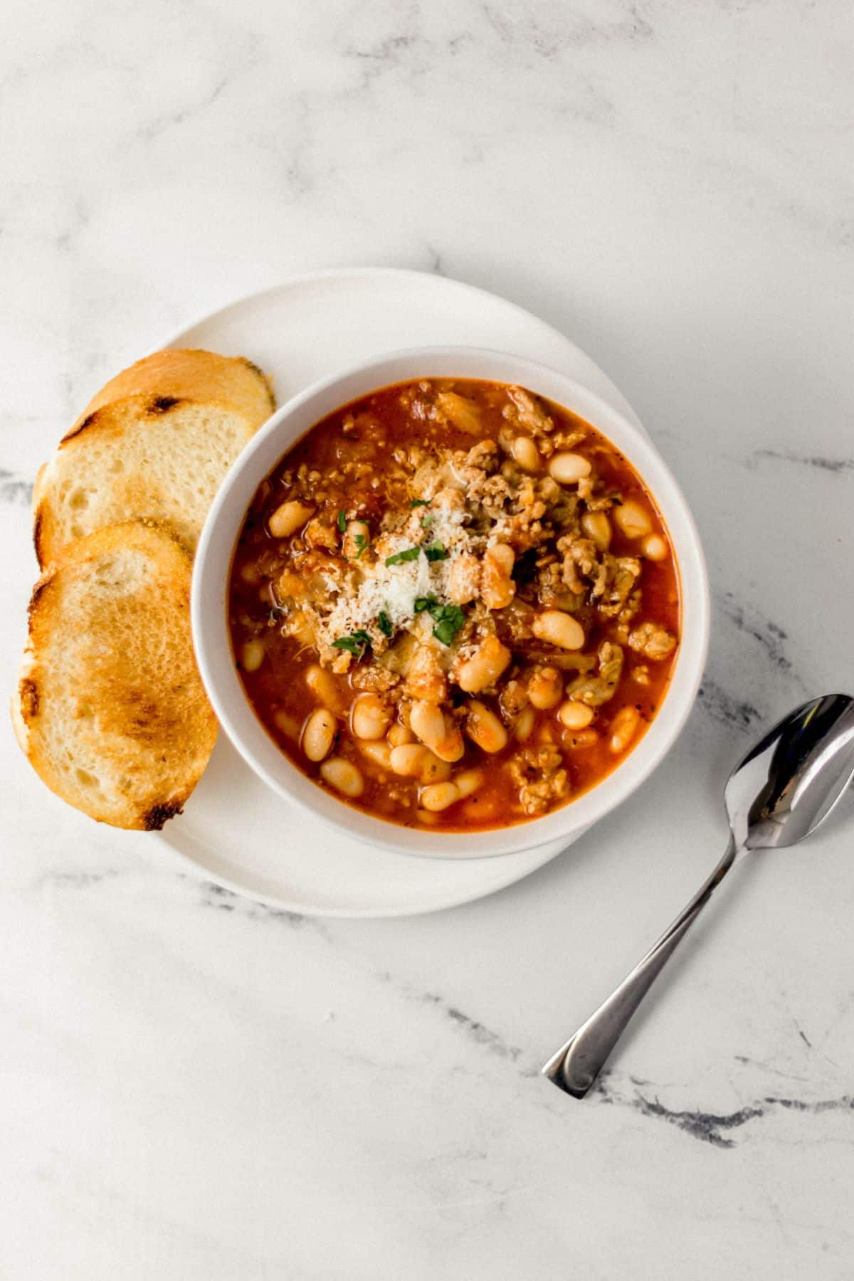 overhead view of bowl of soup over white plate with bread on it beside a spoon