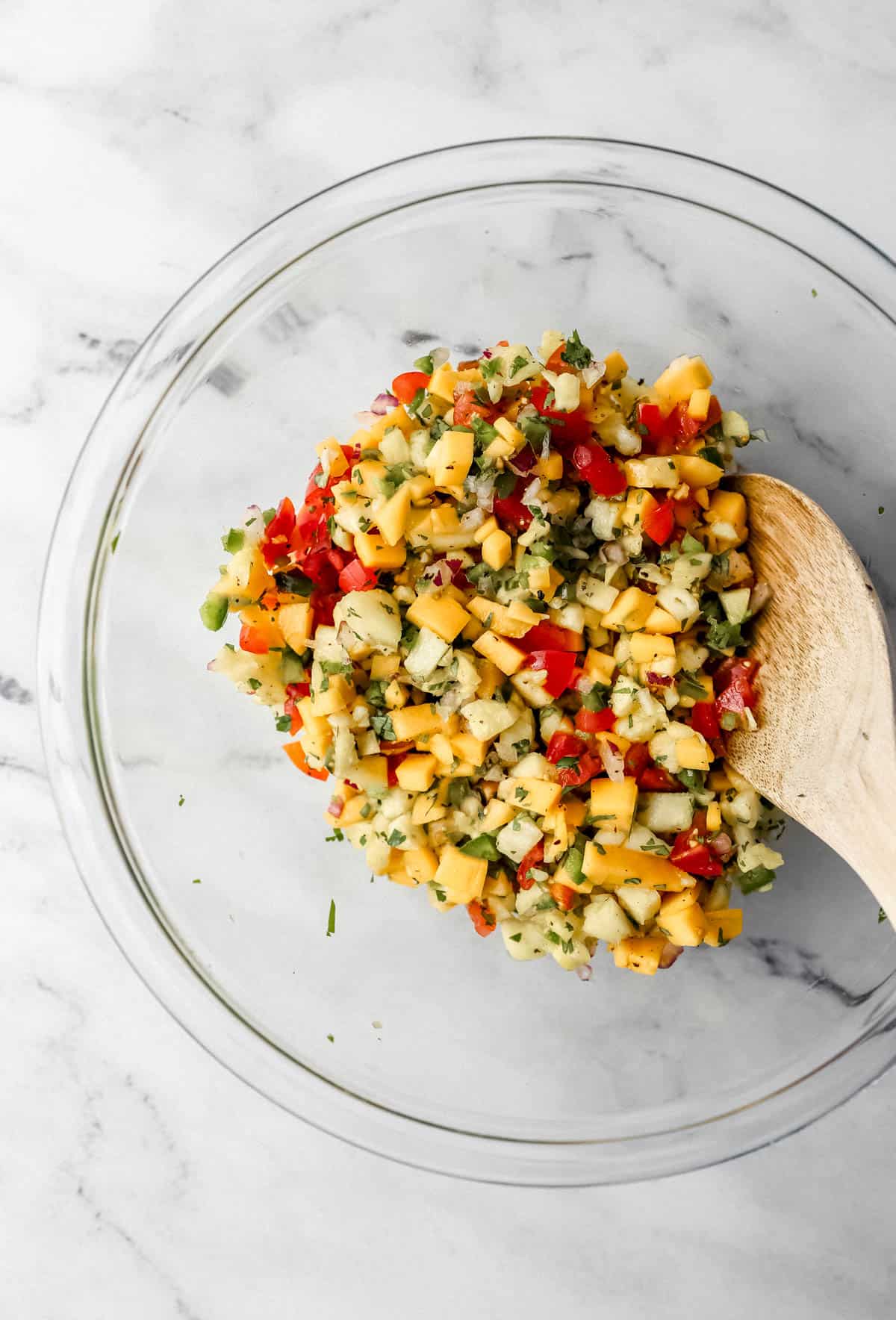 salsa ingredients combined together in glass mixing bowl with wooden spoon 