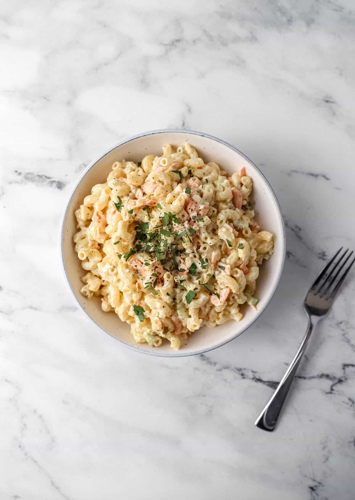 overhead view of pasta salad in white bowl beside a fork on marble surface