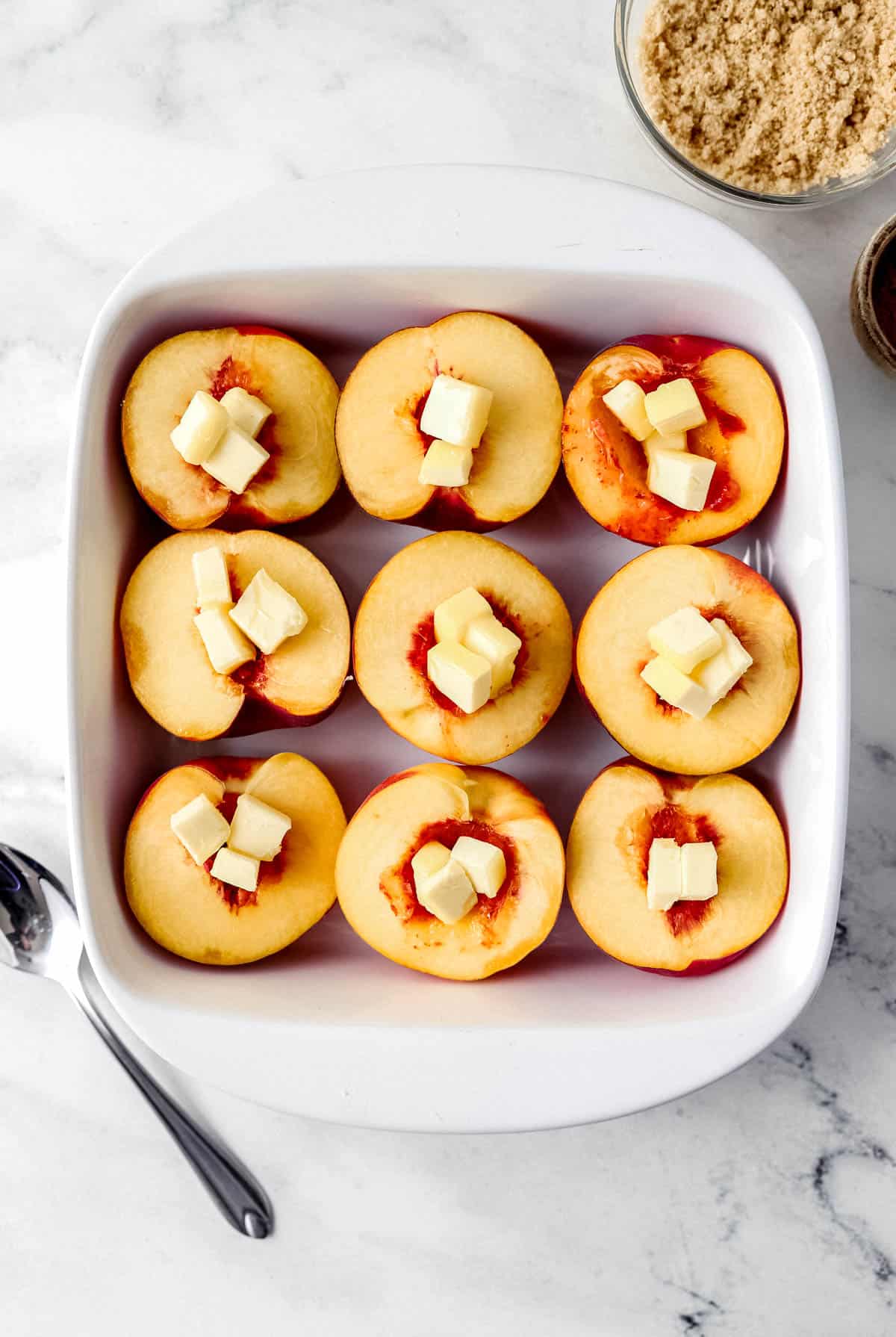 Fresh peach halves topped with cubed butter in white baking dish. 