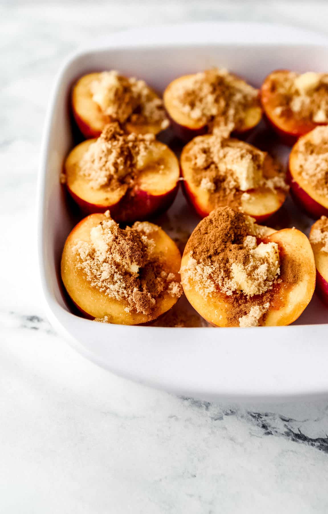 Close up front view of fresh peach halves topped with butter, brown sugar, and cinnamon in white baking dish. 