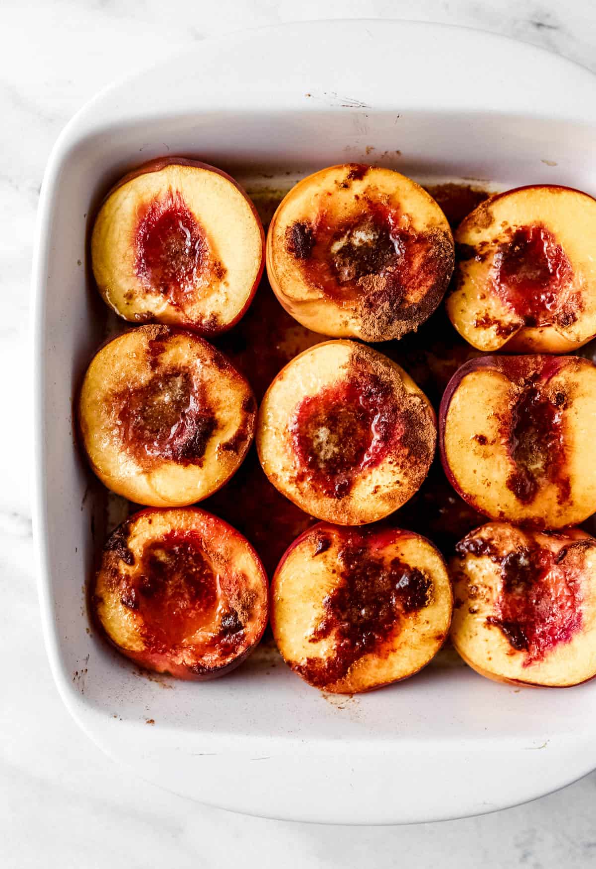 Baked peaches in square white baking dish on marble surface. 