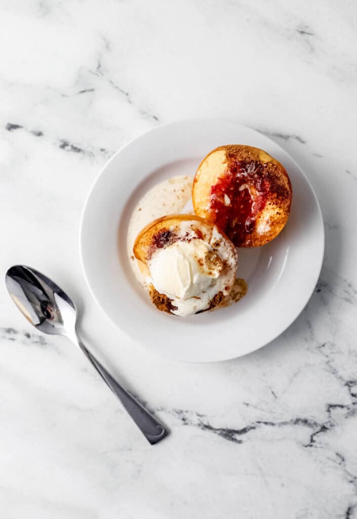 Overhead view of baked peaches on white plate topped with ice cream beside a spoon with text overlay pin.