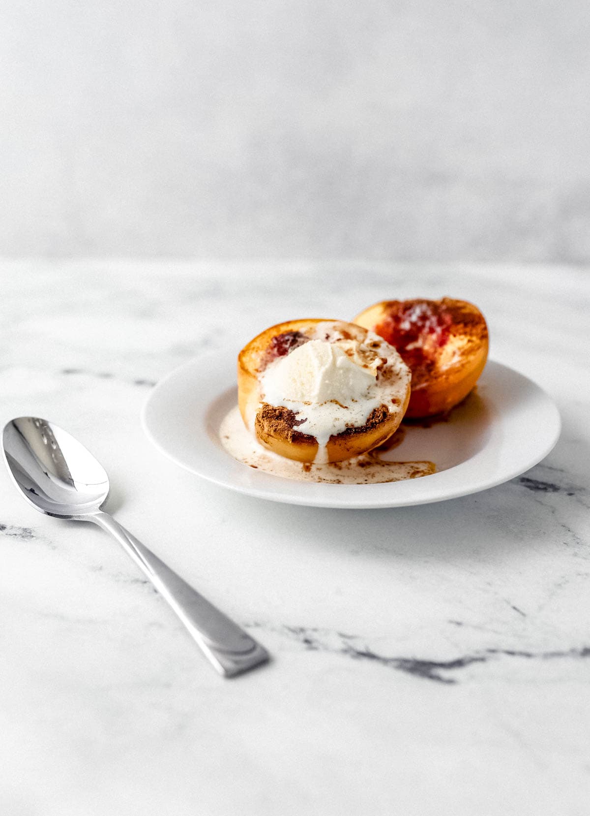 Close up side view of two baked peach halves on white plate topped with ice cream beside a spoon on marble surface. 