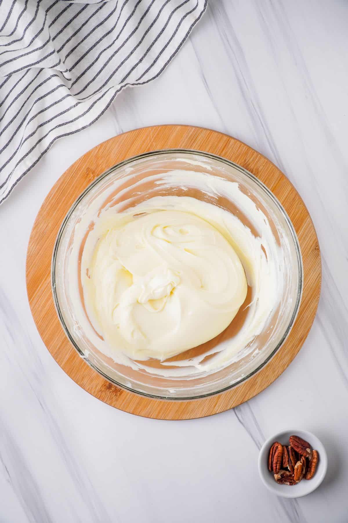 Overhead view of cream cheese, powdered sugar, and whipped topping combined in large glass mixing bowl on round wooden board. 