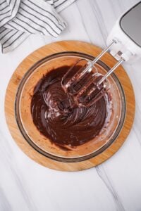 Chocolate pudding mix and milk combined in glass mixing bowl with hand mixer.