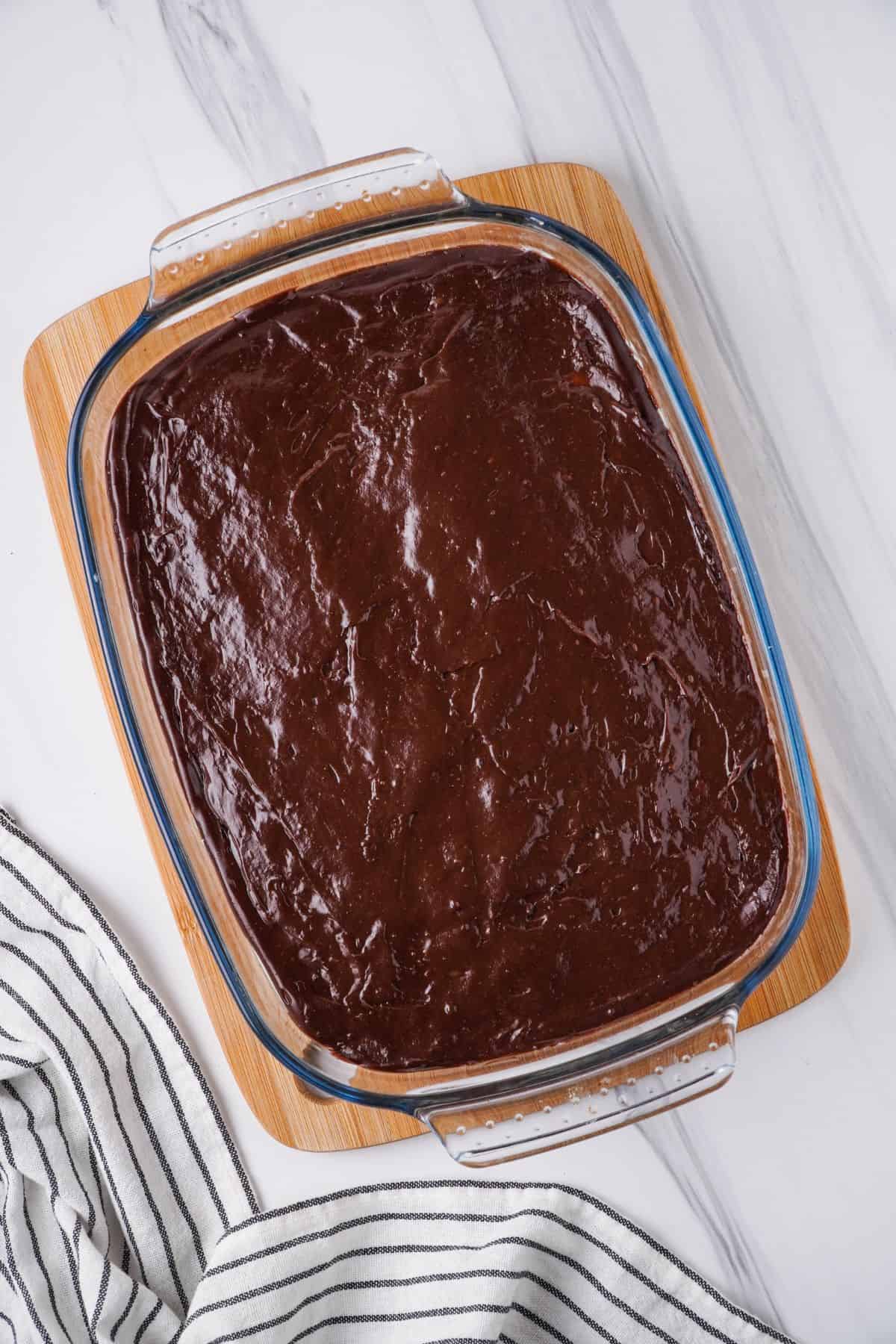 Overhead view of chocolate pudding layer added to glass baking dish over wooden cutting board. 