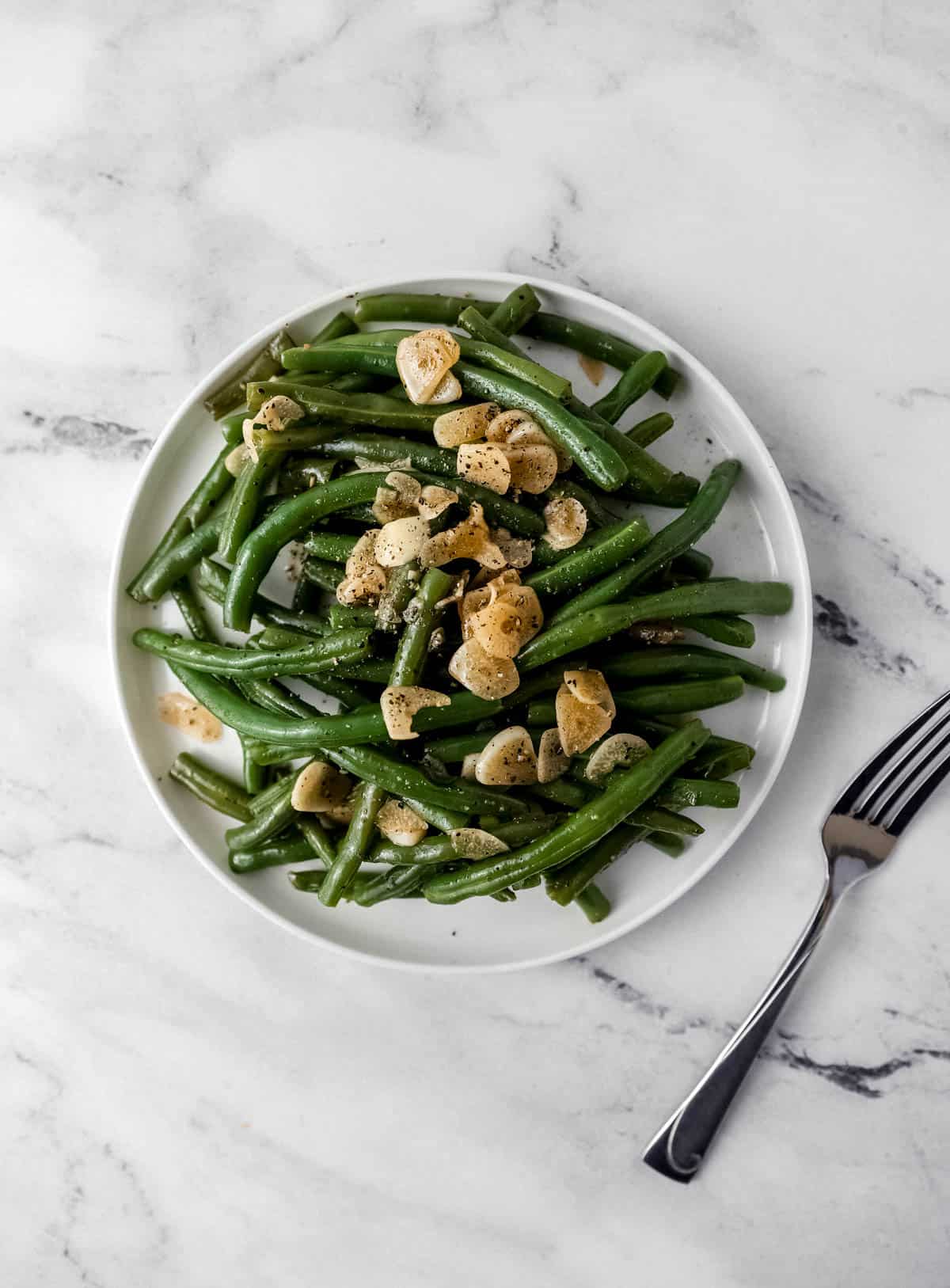overhead view of green beans on white plate beside a fork on marble surface 