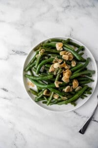 Green beans and garlic added to white plate by fork.