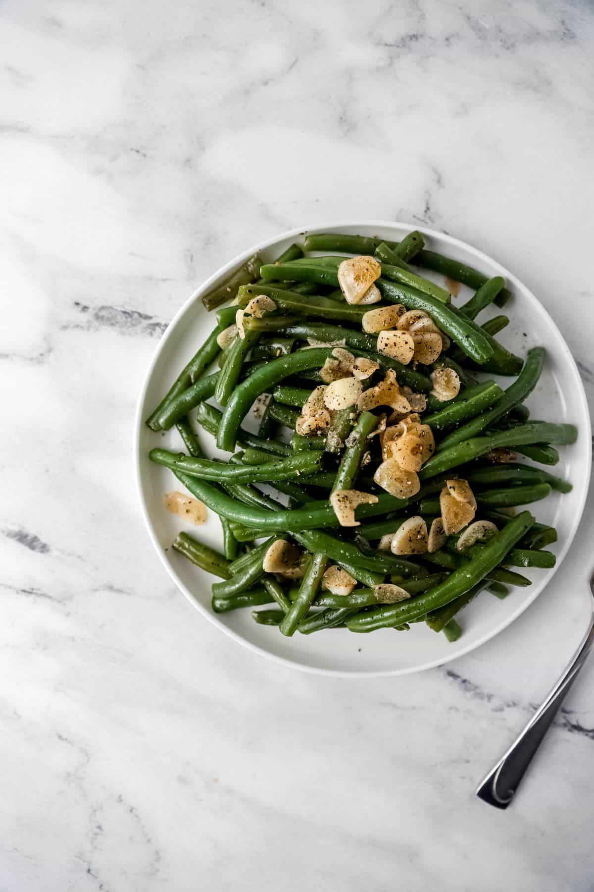 overhead view of finished green beans and garlic on white plate on marble surface