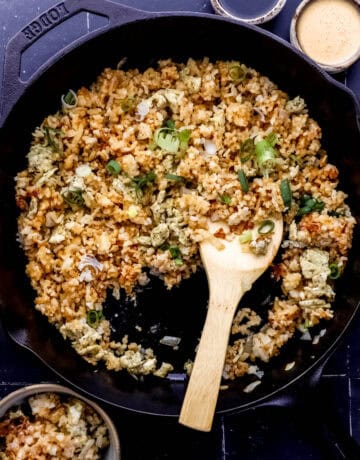 Finished fried rice in cast iron skillet with wooden spoon in it by small bowls of sauce and rice.
