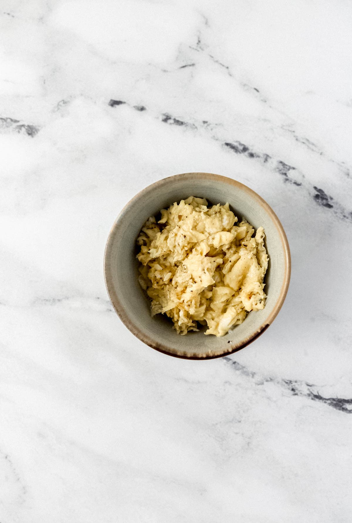 Overhead view of scrambled eggs in small bowl on marble surface.