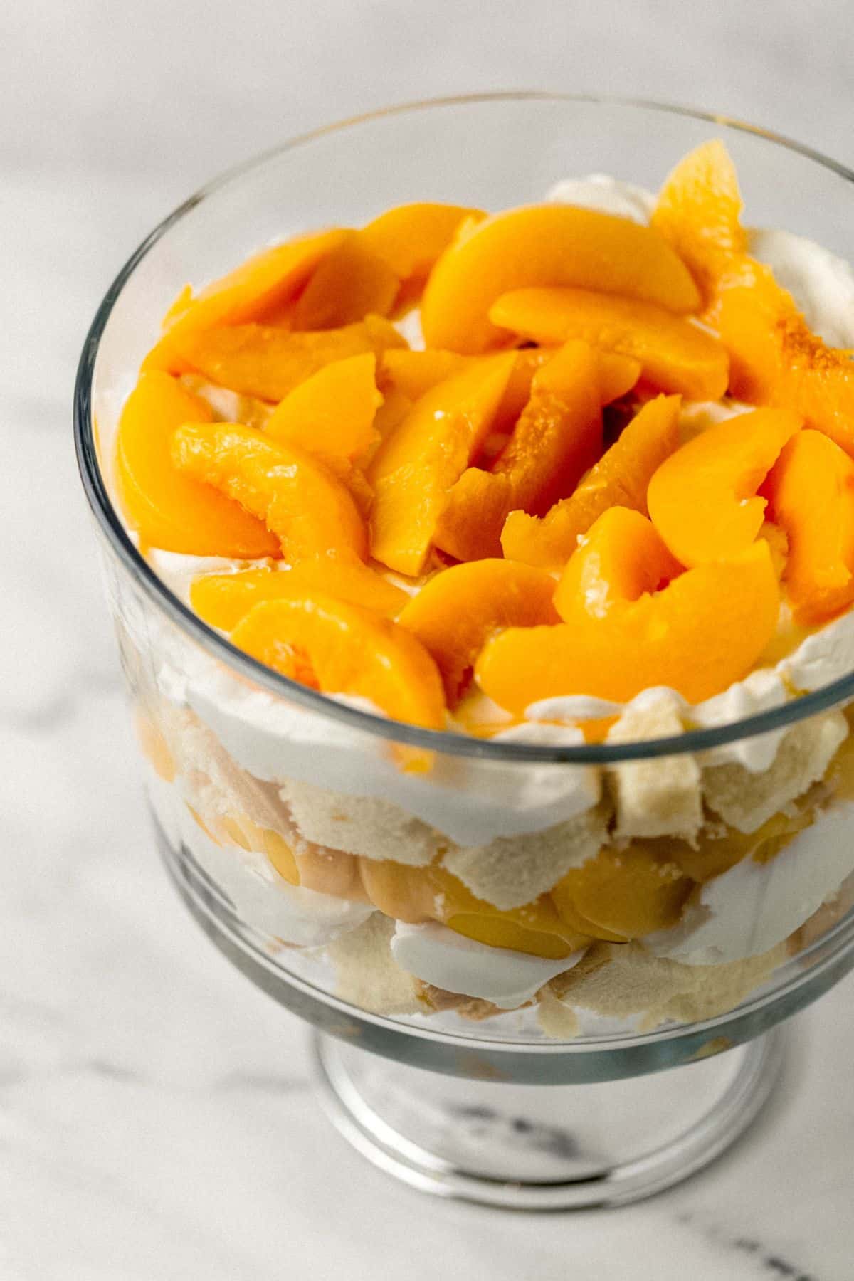 close up view of trifle topped with layer of sliced peaches in glass trifle bowl 