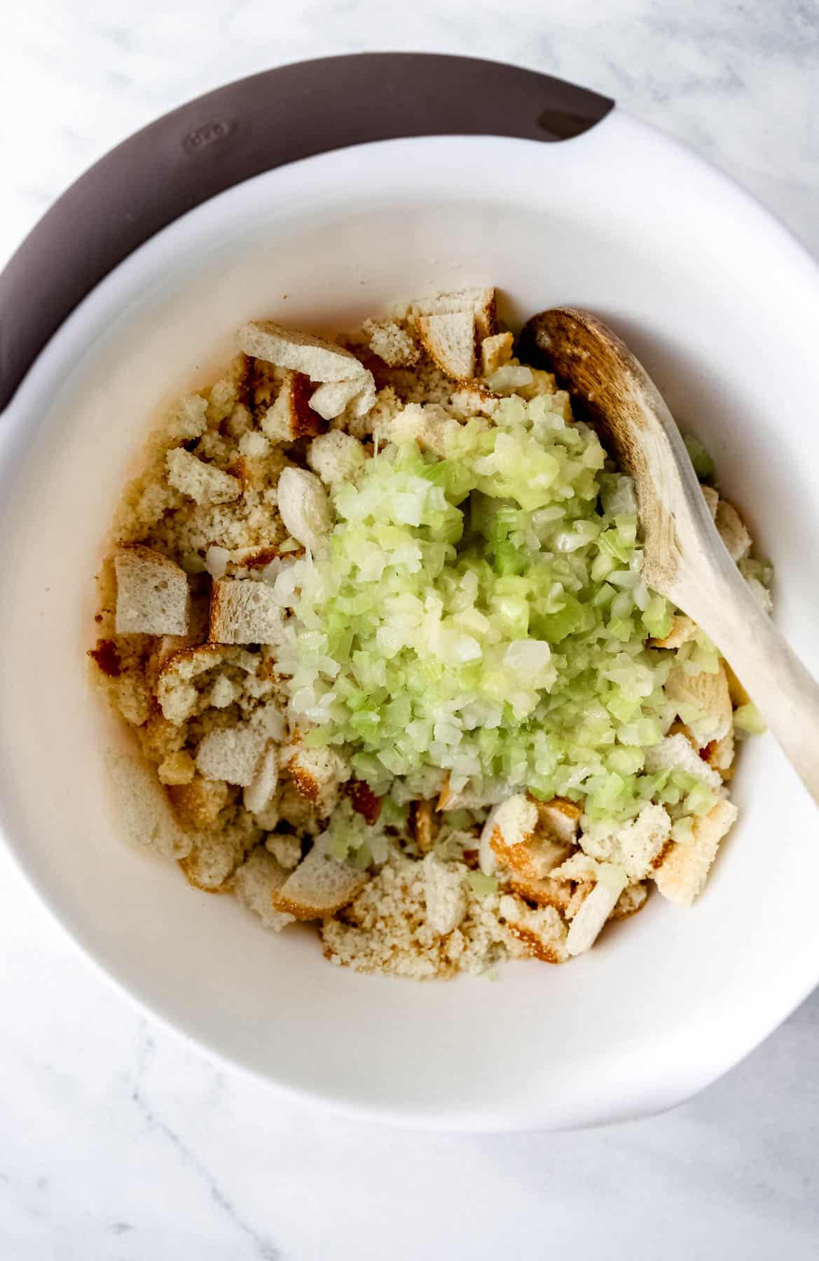 Celery mixture added to bread in large white mixing bowl with wooden spoon. 