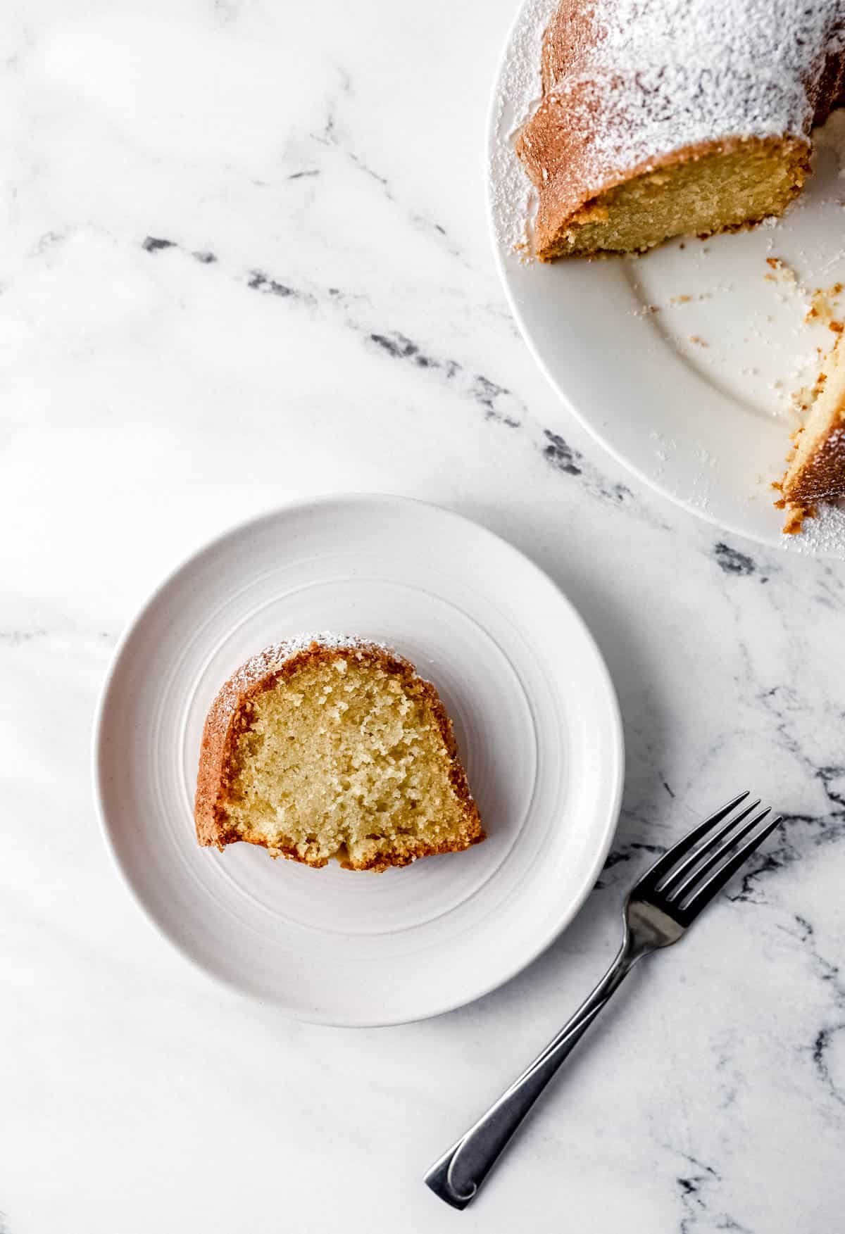 Overhead view of a slice of cake on white plate beside a fork with the rest of the whole cake in the background.