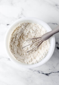 Flour, baking powder, baking soda, and salt in white bowl with whisk.