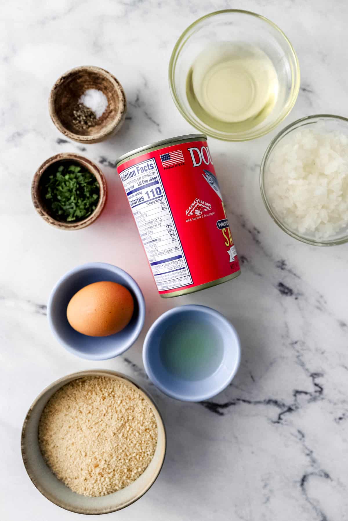 Overhead view of ingredients needed to make salmon patties in separate bowls on marble surface. 