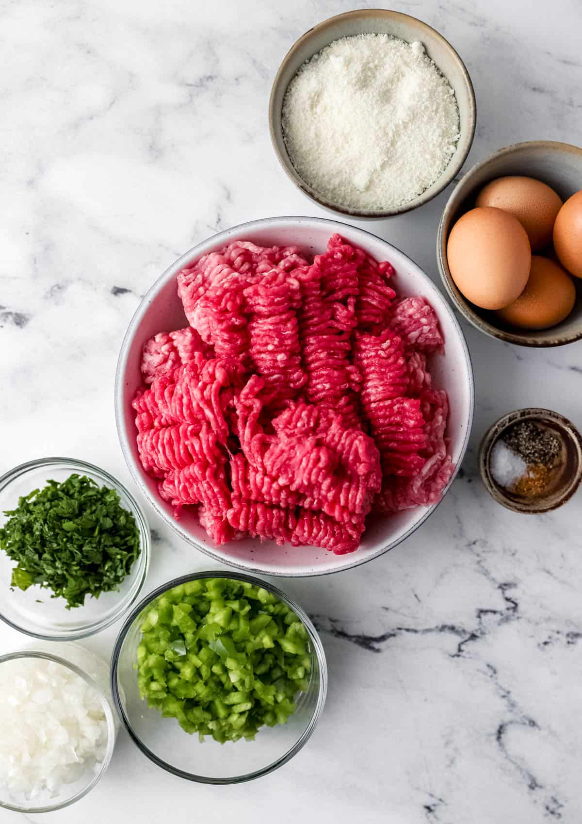 Ingredients needed to make meatloaf in separate bowls on marble surface. 