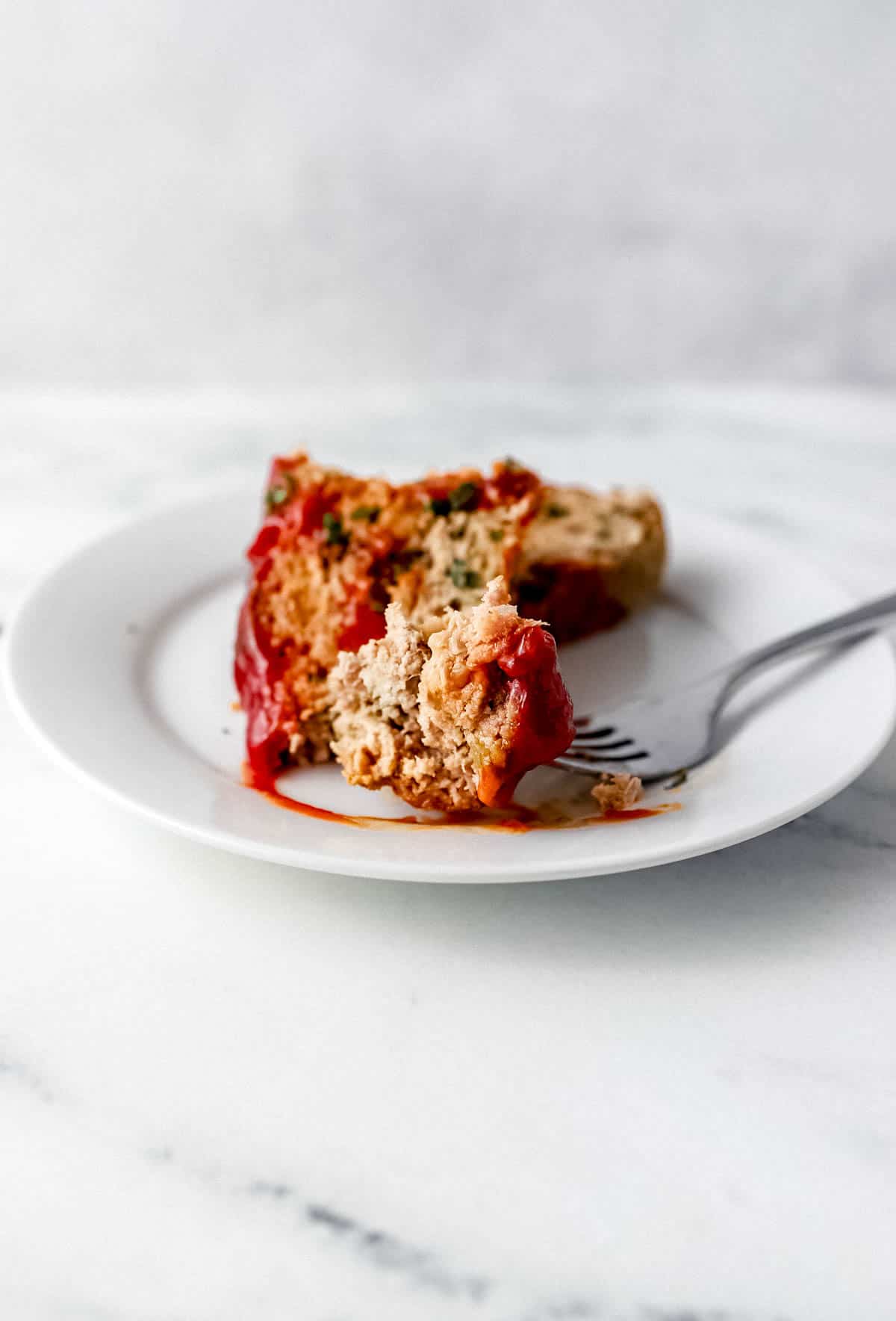 Close up side view of slice of meatloaf on white plate with a fork in it. 