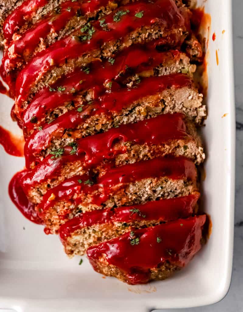 Close up overhead view of sliced meatloaf topped with sauce in white baking dish.