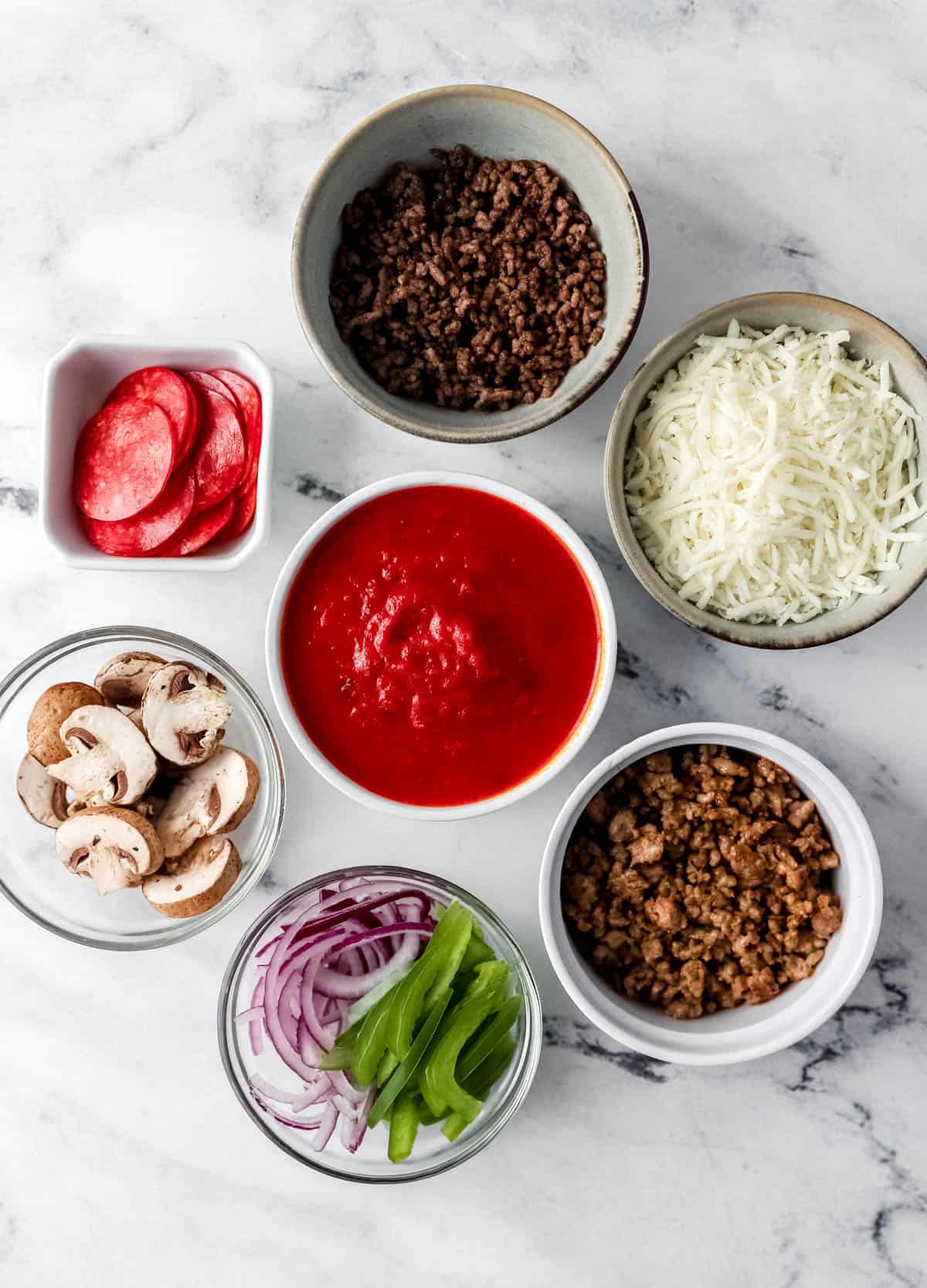 Ingredients needed to make supreme pizza in separate bowls on top of a marble surface. 