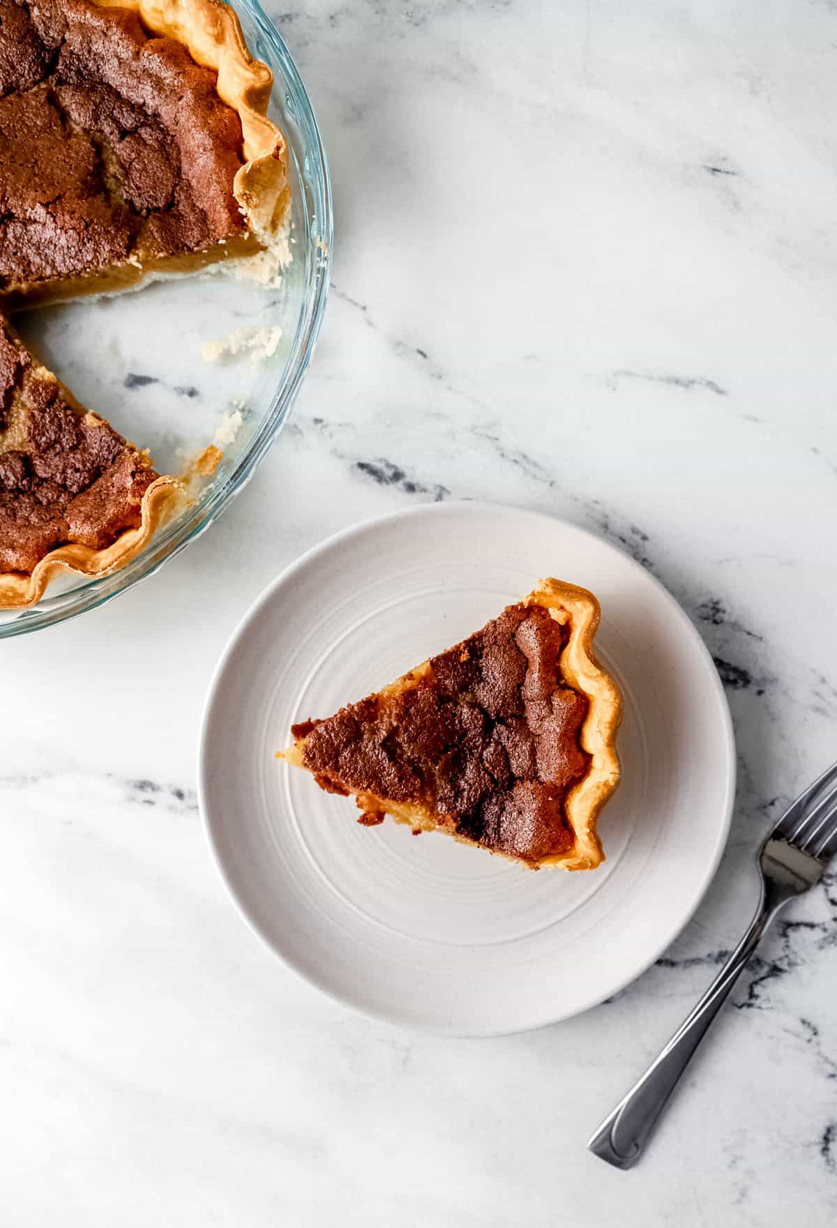 Overhead view of slice of pie on white plate beside a fork with the rest of the pie in the background. 