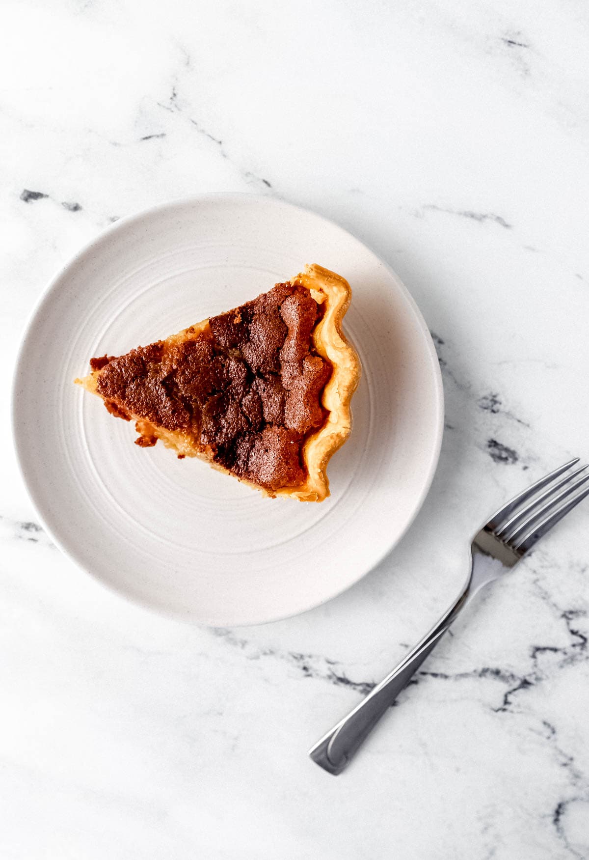 Overhead view of slice of pie on white plate beside a fork on marble surface. 