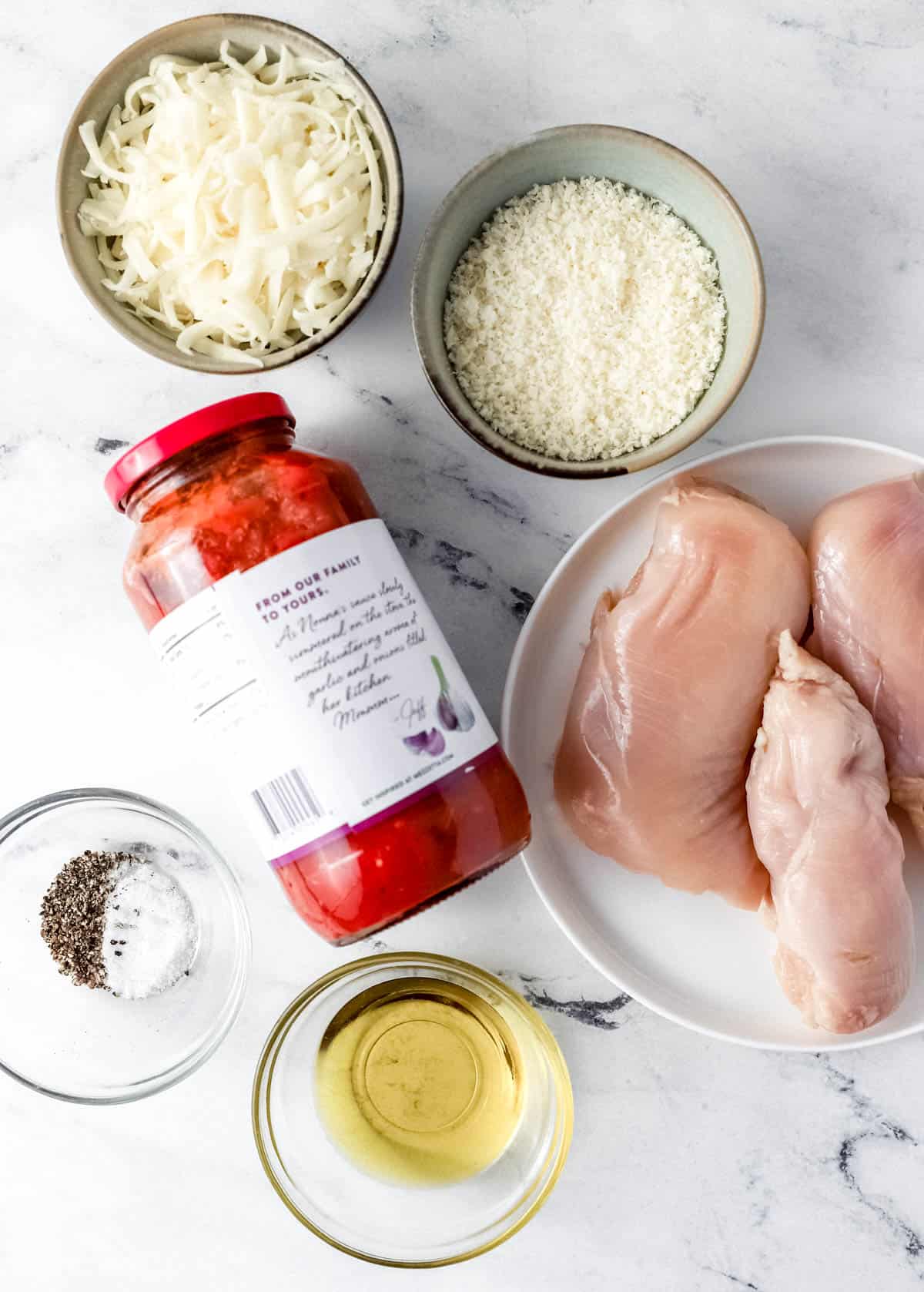 Overhead view of ingredients needed to make chicken mozzarella bake in separate bowls and containers on marble surface. 