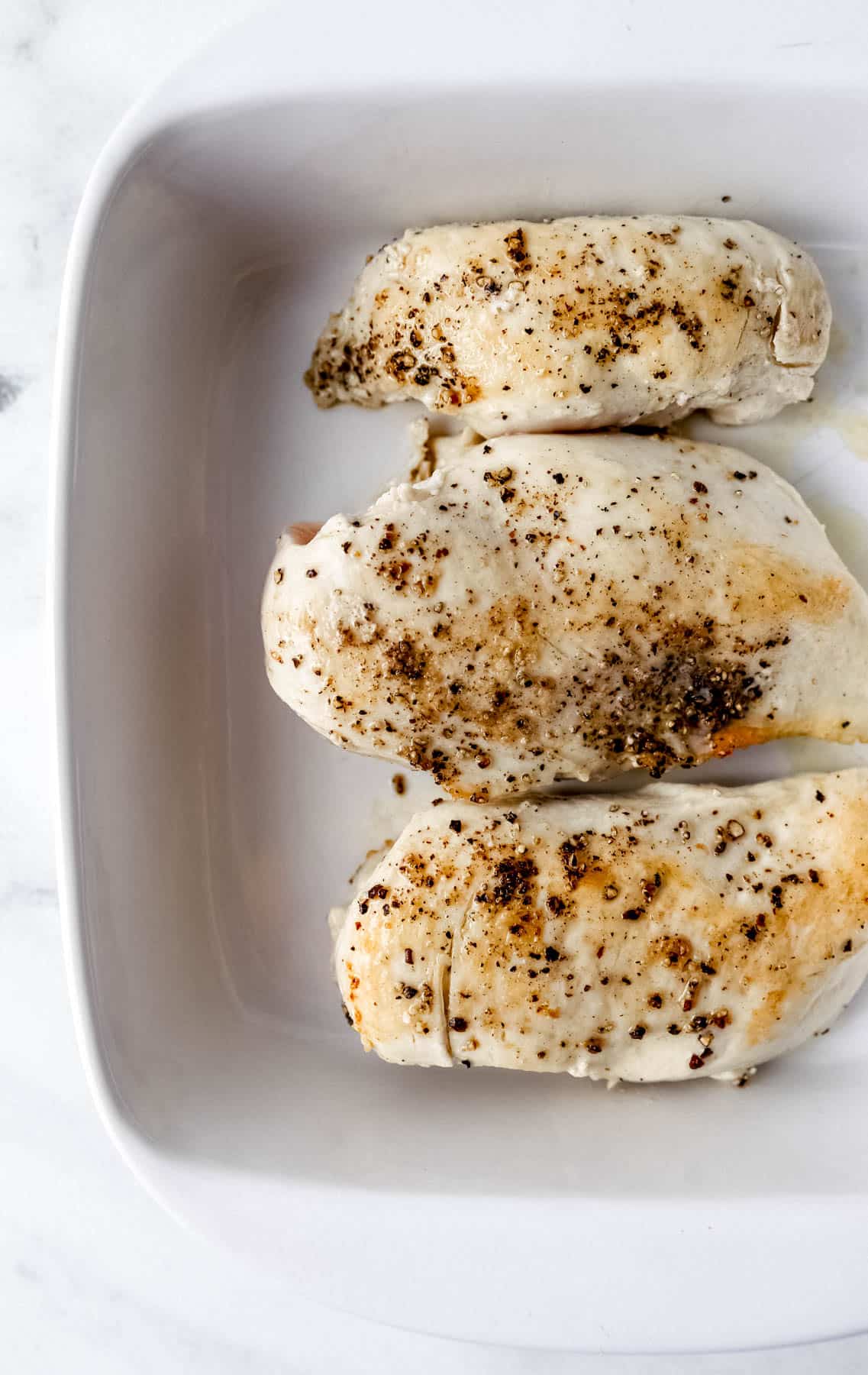 Partially cooked chicken breasts in square white baking dish. 